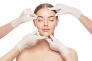 Close up portrait of young woman standing with naked shoulders and neck, face is touched by beauticians in gloves, preparing her for plastic surgery procedures, isolated on white background